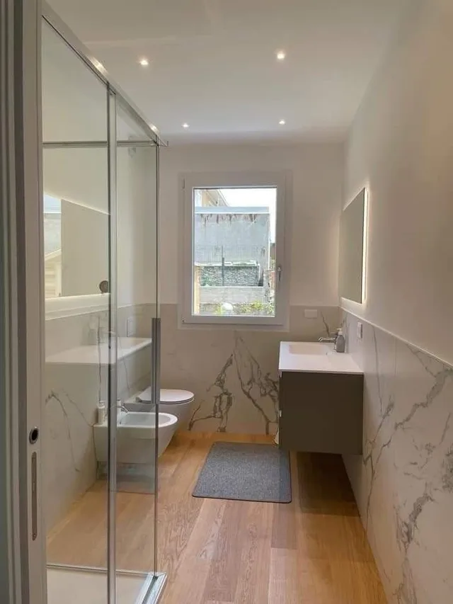 Bagno con parete in vetro, rivestimento effetto marmo e pavimento in legno